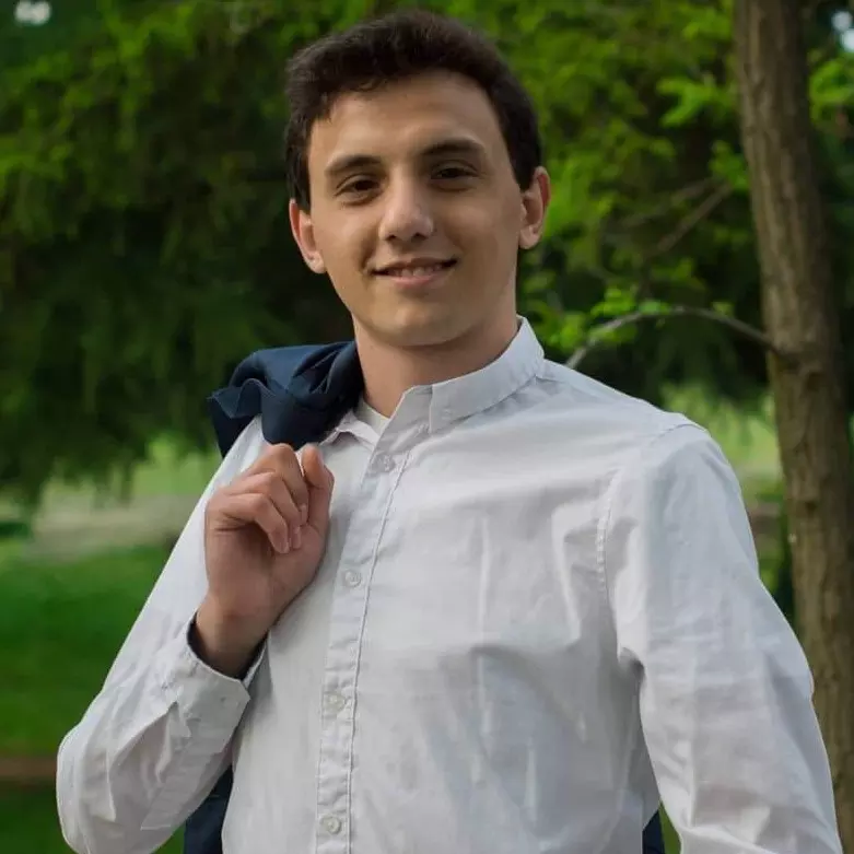 Me, posing infront of greenery. I am smiling, smug. I am wearing a white shirt and holding a blue suit jacket with the pointer finger of my right hand over my right shoulder.